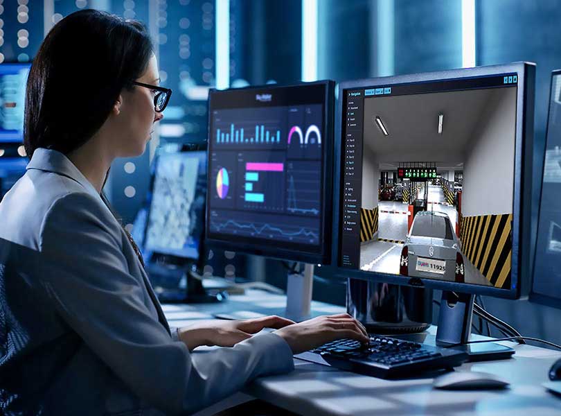 a woman sitting in front of multiple monitors, one showing live feed of a parking area and another displaying log entries.