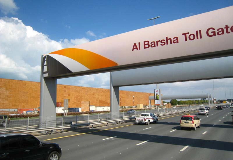 multiple cars driving under a toll gate with a banner reading 'al barsha toll gate,' showcasing automatic toll collection solutions.